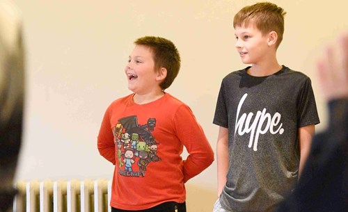 Two young boys stand side by side in a rehearsal or workshop setting. Jack, wearing a red Minecraft t-shirt, laughs joyfully,