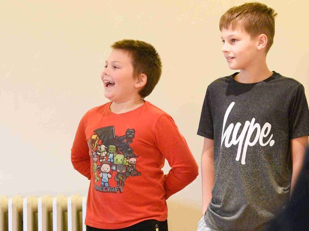 Two young boys stand side by side in a rehearsal or workshop setting. Jack, wearing a red Minecraft t-shirt, laughs joyfully,