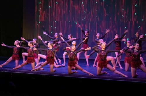 A group of dancers perform on stage in coordinated poses, wearing red and black sequined costumes with fringe skirts, arms extended in expressive movement.