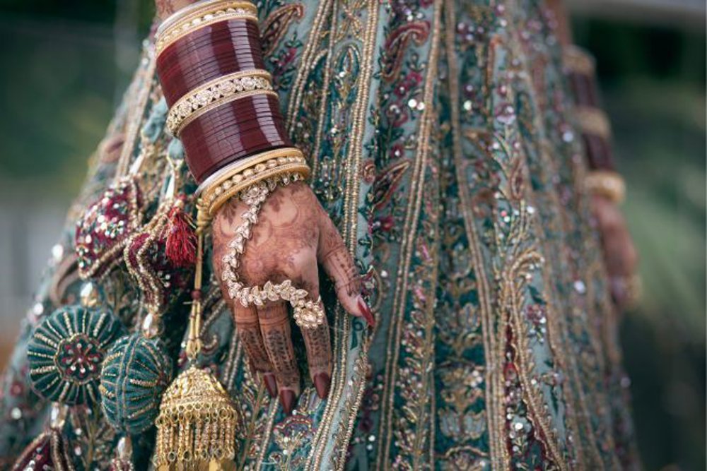 A close-up of a hand adorned with intricate henna designs, gold jewellery, and red bangles, resting against a heavily embroidered and sequined lehenga in shades of green and gold. Decorative tassels and gold accents enhance the traditional bridal attire