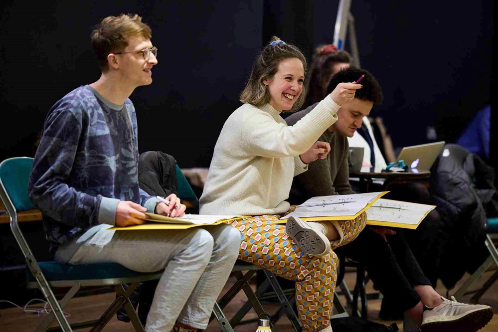 A rehearsal room with three people sitting on chairs. One is wearing a patterned sweatshirt and holding a notebook, another is smiling and gesturing while wearing a cream jumper and colourful patterned trousers, and a third person is seated, focusing on a script.