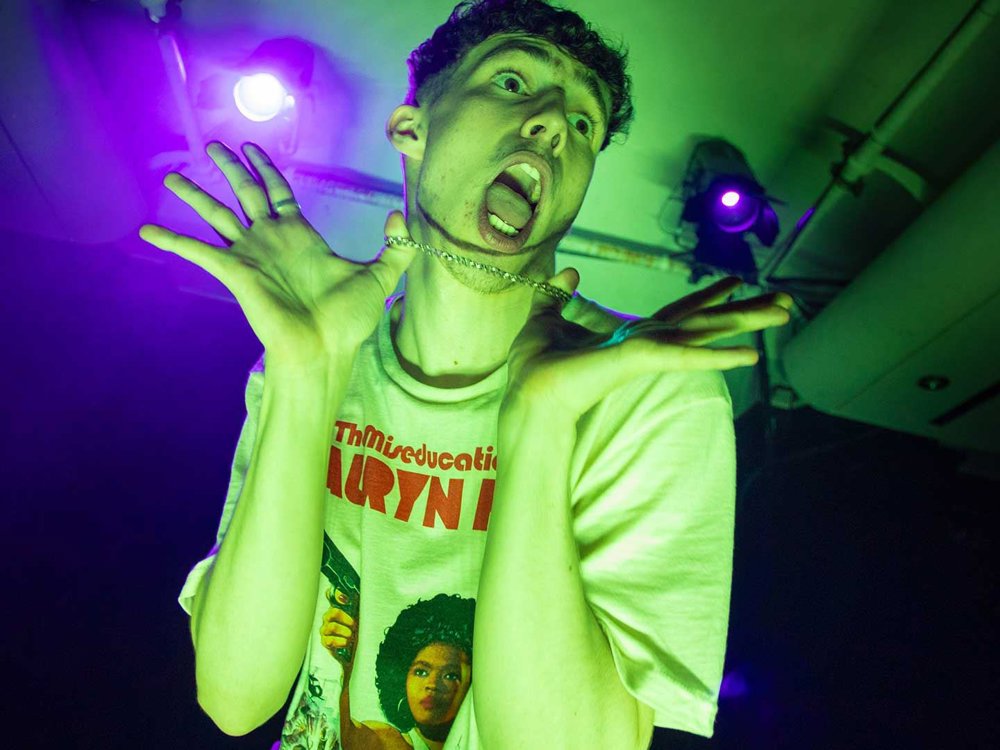 A young man is making a playful and exaggerated expression, holding his necklace with both hands. He is wearing a T-shirt that reads 'The Miseducation of Lauryn Hill' and is bathed in green stage lighting.