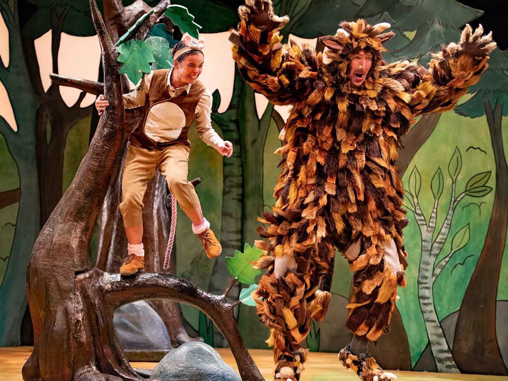 A large, shaggy creature leaps with raised arms on a woodland stage, while a small mouse-like character balances on a tree branch nearby, appearing playful and energetic.