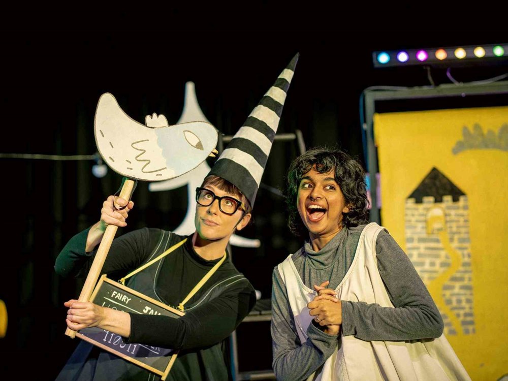 Rapunzel and the witch pose playfully together on stage. The witch holds a bird-shaped prop and a sign, while a painted tower backdrop and colourful set pieces frame the scene.