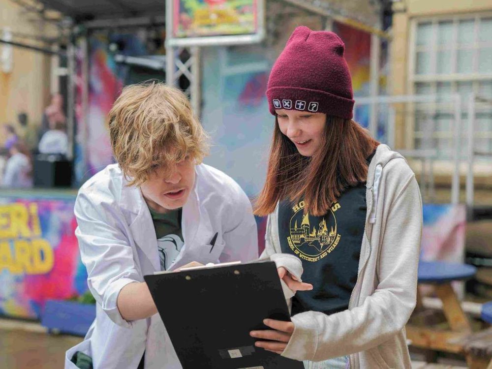 Two teenagers engage with a clipboard in an outdoor setting. One wears a white lab coat and gestures towards the clipboard, while the other, dressed in a hoodie and maroon beanie, smiles while looking at it. A colourful 'SUMMER COURTYARD' sign and blurred background activity suggest a creative or educational event.