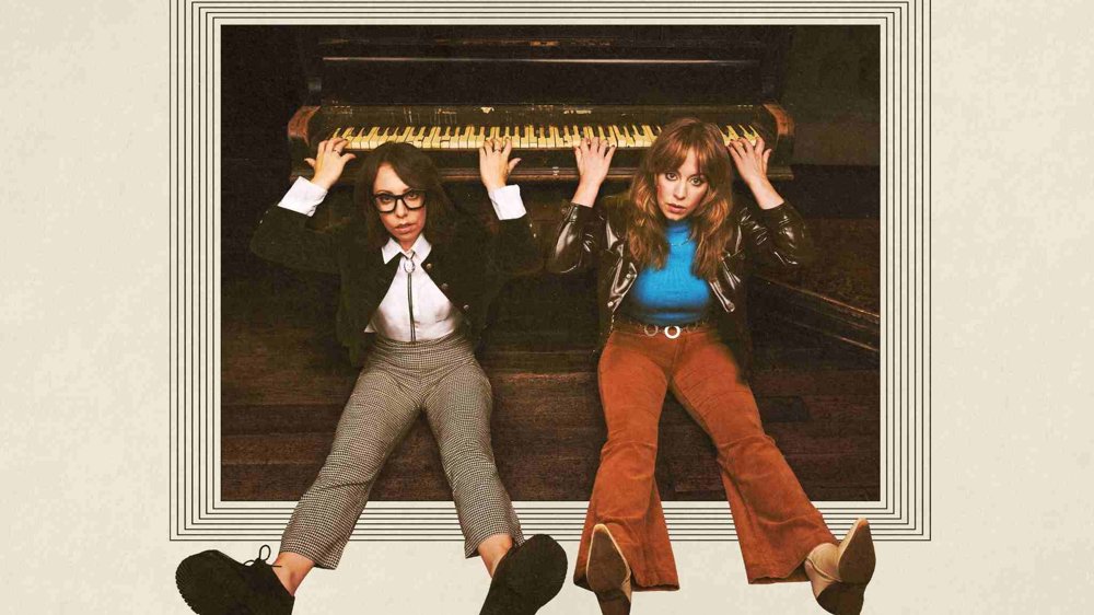 Two people sit on the floor beneath an upright piano, holding onto the piano keys above them. They look directly at the camera with serious expressions, wearing retro-style clothing, framed by a graphic border that gives the image a vintage feel.