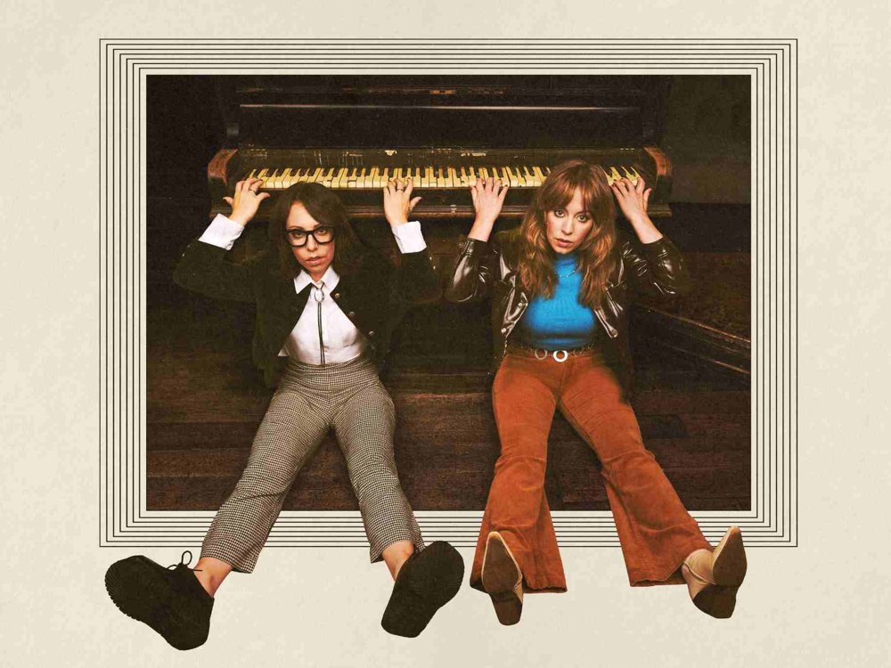 Two people sit on the floor beneath an upright piano, holding onto the piano keys above them. They look directly at the camera with serious expressions, wearing retro-style clothing, framed by a graphic border that gives the image a vintage feel.