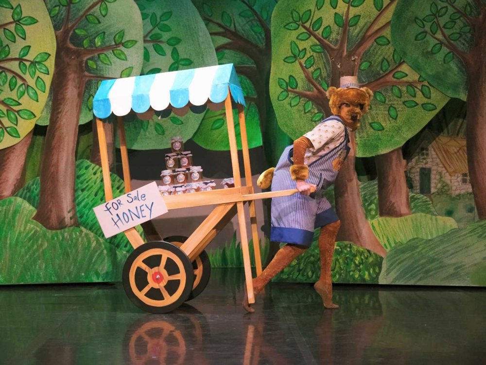A performer dressed as a bear in striped overalls pulls a wooden cart filled with honey jars, set against a painted forest backdrop.