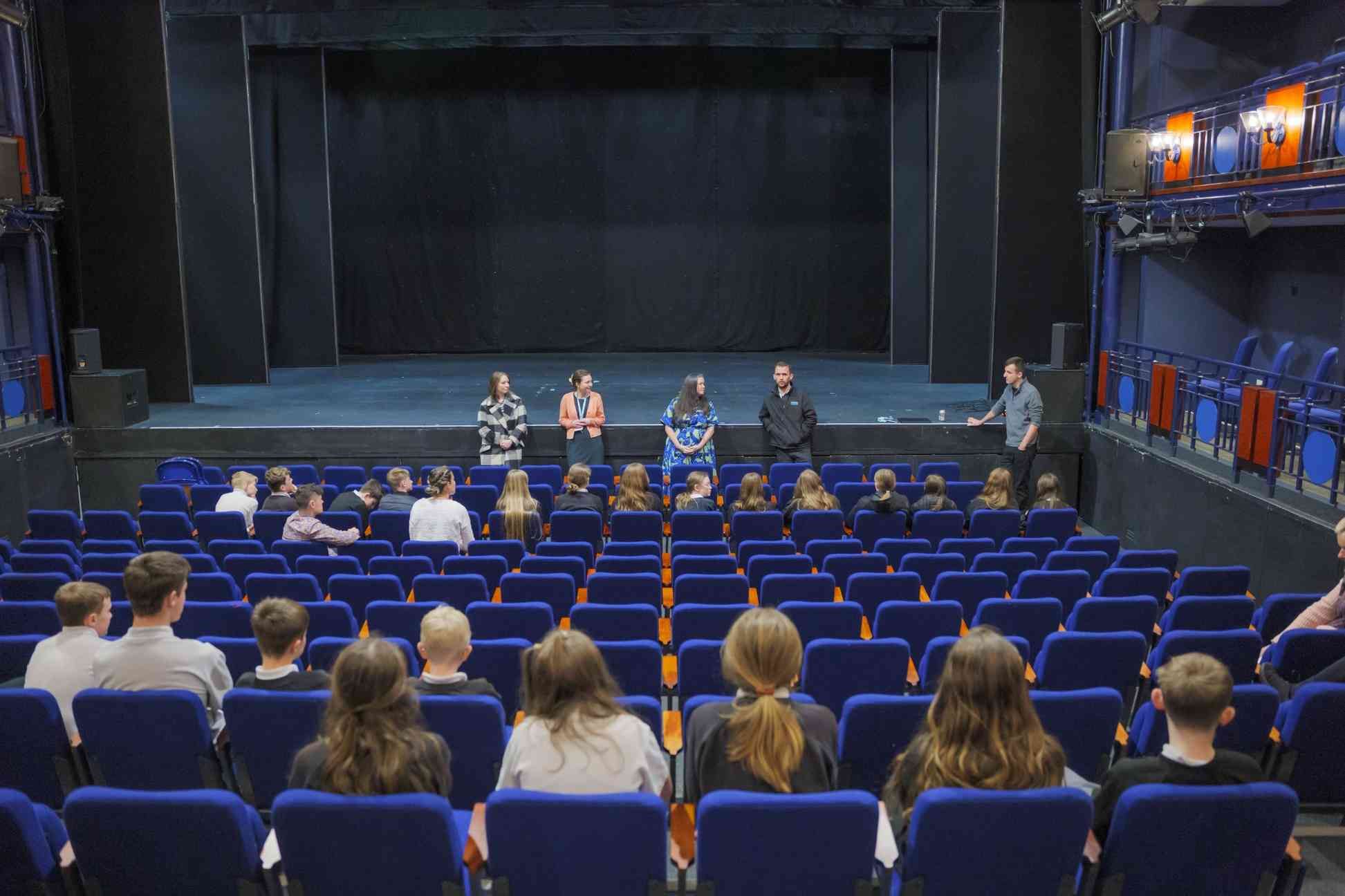 Students seated in a theatre auditorium listening to staff standing at the front of the stage during a school visit or workshop.