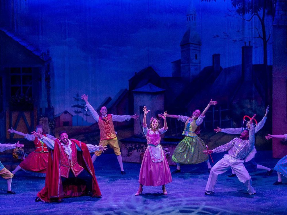 A full company dance number on stage, with performers in colourful village costumes striking joyful poses beneath blue theatrical lighting, suggesting a lively opening scene in a fairytale village.