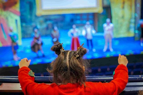 A young girl wearing a festive red jumper with reindeer hair clips watches a colourful stage performance at Lawrence Batley Theatre, raising her arms in excitement from the balcony.