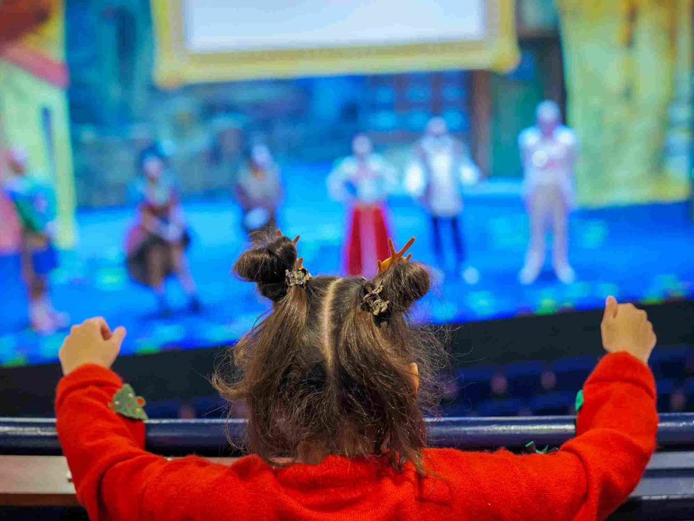 A young girl wearing a festive red jumper with reindeer hair clips watches a colourful stage performance at Lawrence Batley Theatre, raising her arms in excitement from the balcony.