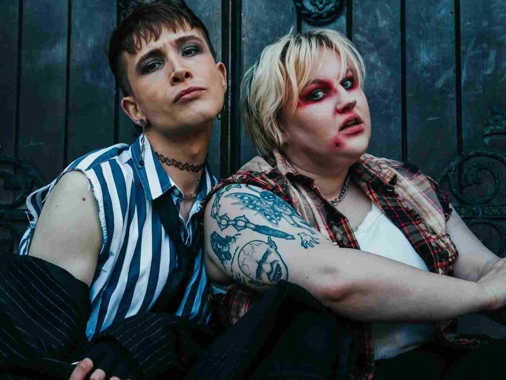 Two people sit close together against a dark wooden door with ornate metalwork. One wears a striped shirt and dark eye makeup, while the other has red eye makeup, a checked shirt, and visible tattoos.