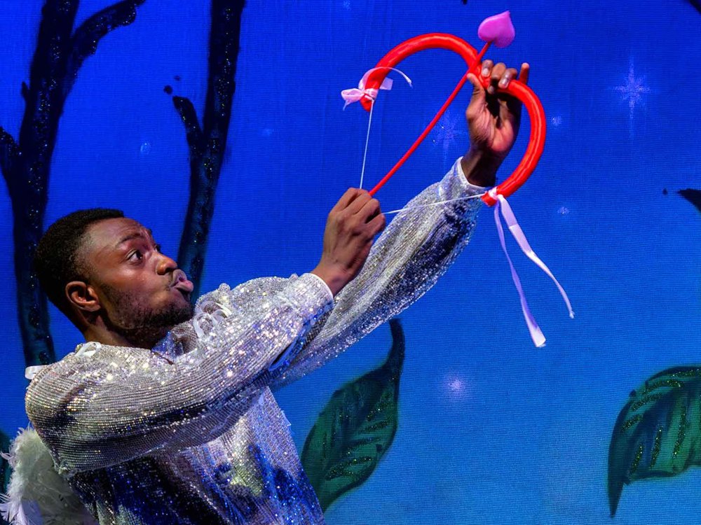 A performer in a sparkling costume holds up a a red bow with a small heart shaped arrow, lit by rich blue stage lighting that creates a magical, romantic atmosphere.