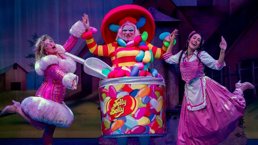Three characters perform centre stage: a flamboyantly dressed character bursting out of an oversized jelly bean tin, flanked by two excited performers in pink costumes, creating a bright, comic moment.