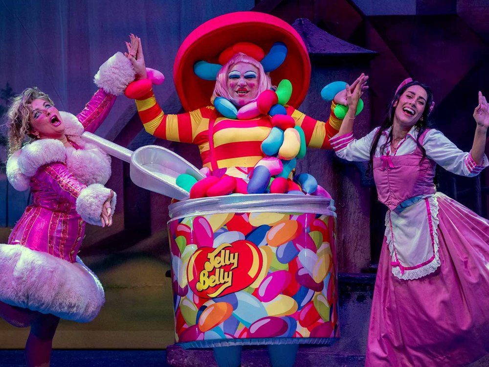 Three characters perform centre stage: a flamboyantly dressed character bursting out of an oversized jelly bean tin, flanked by two excited performers in pink costumes, creating a bright, comic moment.