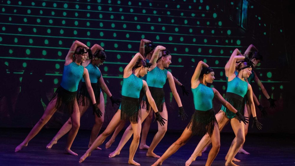A group of dancers perform on stage in matching turquoise and black fringed costumes, striking synchronized poses against a backdrop of glowing green dots.