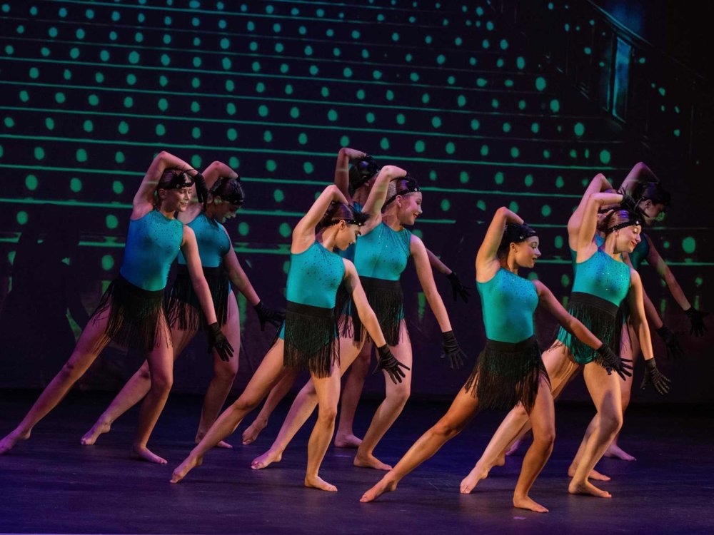 A group of dancers perform on stage in matching turquoise and black fringed costumes, striking synchronized poses against a backdrop of glowing green dots.