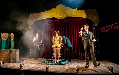 A magic trick is happening in the centre of the stage where a person wearing a gold outfit has six silver hoops around her legs from her feet to her waist. 