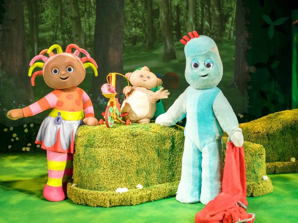 Upsy Daisy, Makky Wakkadoo and Igglepiggle pose together on stage in a forest setting, smiling and waving beside Makky’s tricycle.