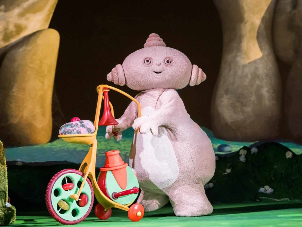 Makky Wakkadoo pushes her colourful tricycle on stage, smiling in front of a rocky cave setting in In the Night Garden… LIVE!