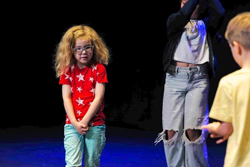 A young girl stands centre stage during an Introduction Stage class, wearing a red t-shirt with white stars and light blue trousers. She looks slightly shy or thoughtful.