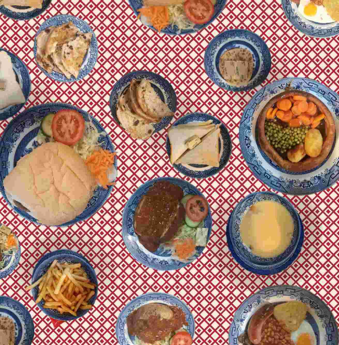 A colourful overhead pattern of blue plates filled with classic café foods—including sandwiches, soup, fries, roast dinners, vegetables and desserts—arranged on a red and white checkered tablecloth.