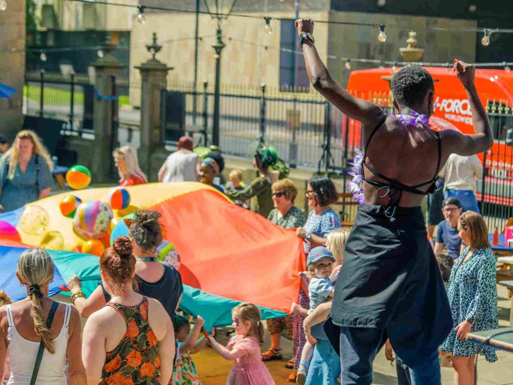 A lively outdoor festival scene with a crowd gathered around a large, multicoloured parachute filled with beach balls. A performer dressed in black, wearing a purple lei, raises their arms in excitement while facing the crowd. String lights and a red van are visible in the background.