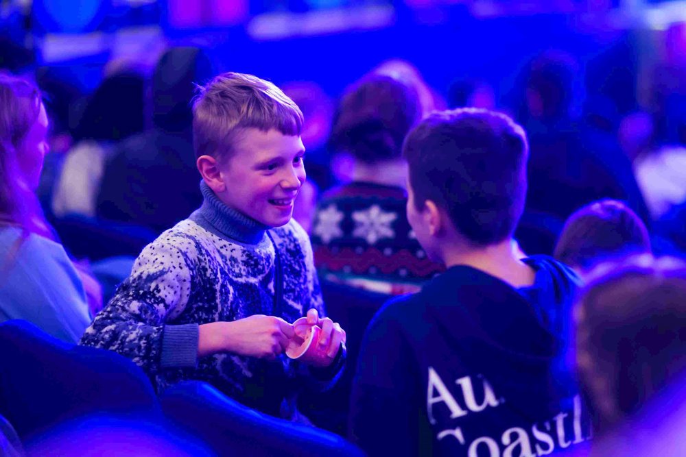 Two boys seated in a theatre audience, one wearing a patterned jumper and smiling warmly while holding a snack, engaging in conversation with the other. 