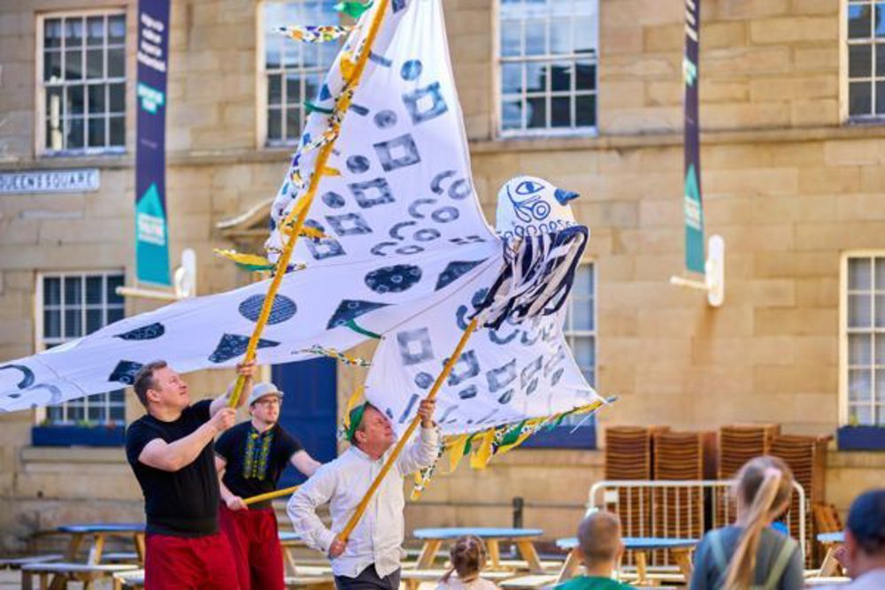 Three men are outdoors holding large, decorative bird-shaped puppets on poles, performing in a courtyard area. The fabric wings feature bold, black patterns, and people are watching the performance in the foreground. A stone building with large windows and event banners forms the backdrop.