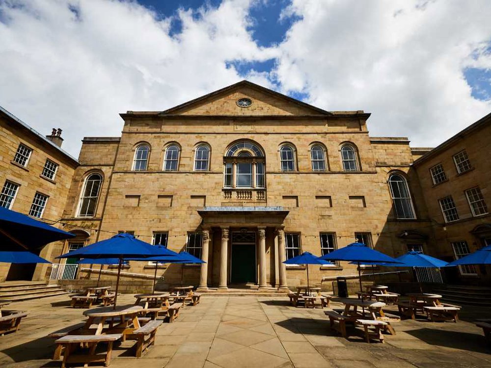 Picture of Lawrence Batley Theatre, which is a Georgian stone building set back behind a courtyard with wooden tables and benches, with blue umbrellas, under a blue cloudy sky in Huddersfield