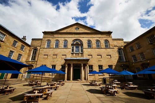 Picture of Lawrence Batley Theatre, which is a Georgian stone building set back behind a courtyard with wooden tables and benches, with blue umbrellas, under a blue cloudy sky in Huddersfield