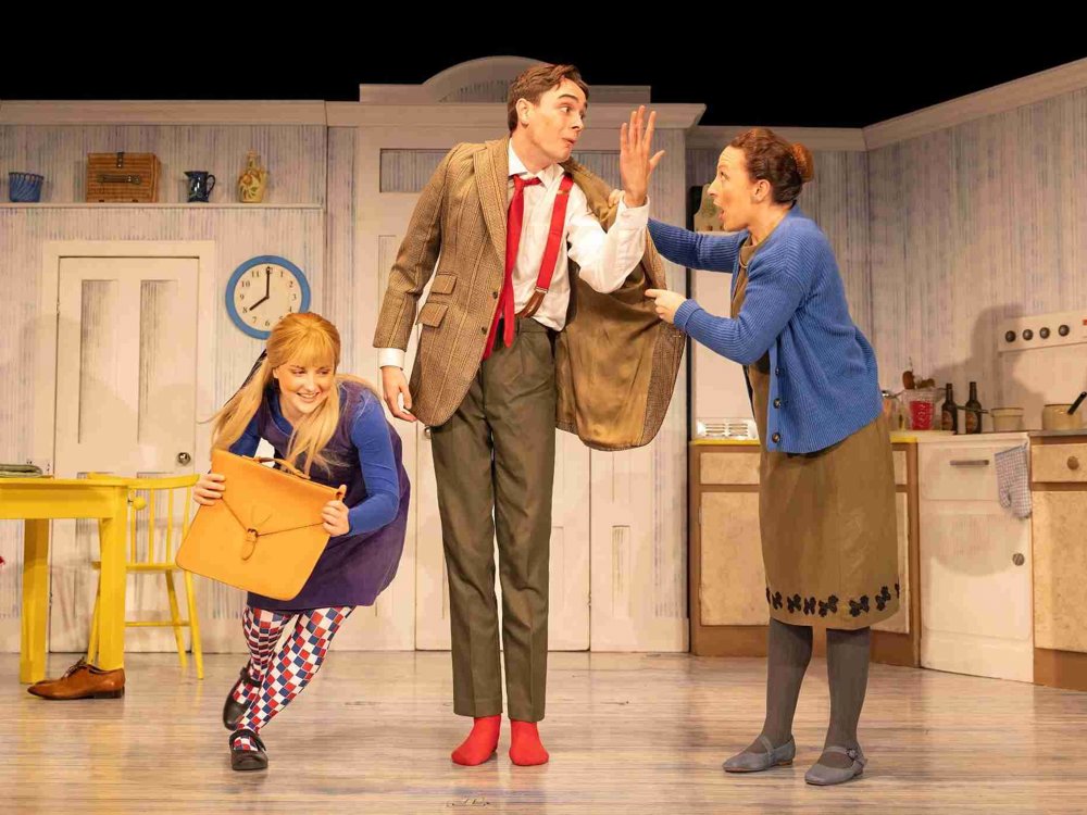 In a brightly lit kitchen set, a young woman crouches with a large envelope while a tall man in a jacket and red socks reacts dramatically, and another woman points at him in surprise.
