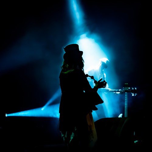 Moody stage scene with a silhouetted musician in a top hat playing percussion in the foreground, lit by blue spotlight beams, with another musician faintly visible in the background.