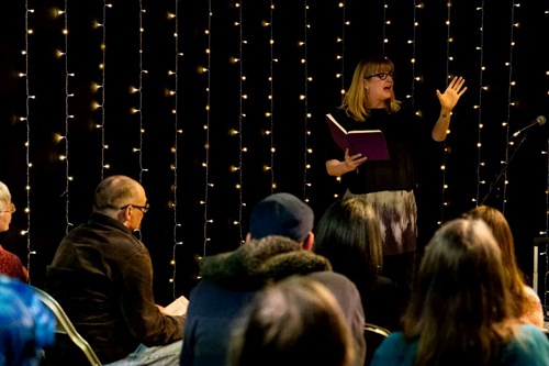 Host of Attic Stories, Rose Condo, stands in front of a seated crowd reading from an open book. Behind her, there is a backdrop of fairy lights against a black curtain. 