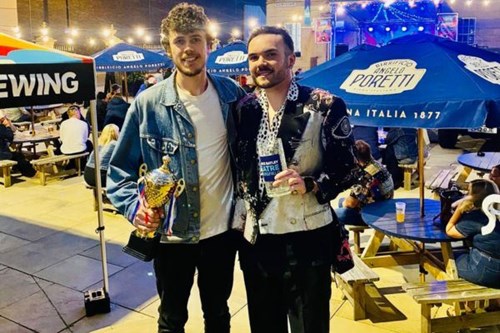 Two men pose outside on stone steps at night, smiling and holding trophies. One wears a denim jacket and holds a gold trophy, while the other wears a patterned shirt and holds a glass engraved with "Lawrence Batley Theatre". People sit at outdoor benches under branded parasols in the background.