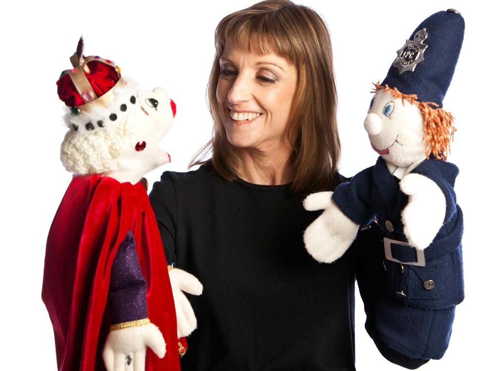 A smiling woman dressed in black holds two hand puppets against a plain white background. One puppet is dressed as a king with a red velvet cape and crown, while the other is a police officer in a dark blue uniform and tall helmet. The woman looks warmly toward the puppets as if bringing them to life in conversation.