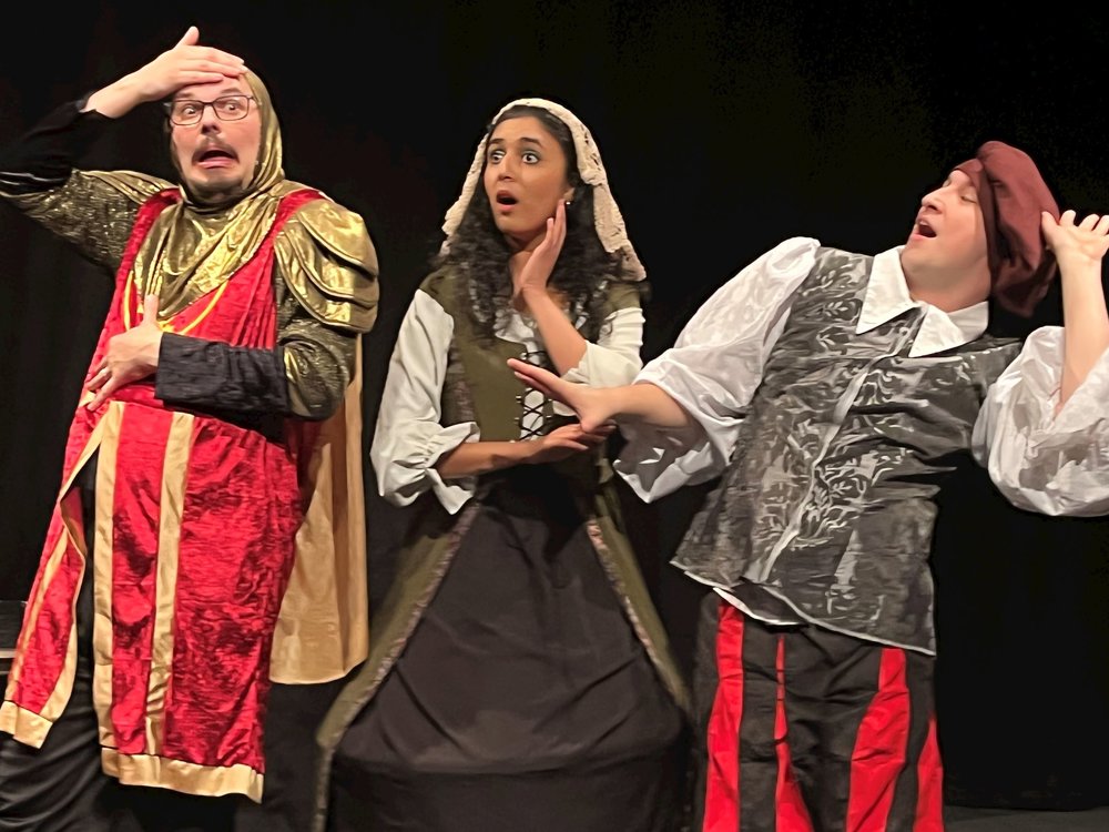 Three performers react dramatically in a comic scene, wearing colourful historical costumes and striking surprised poses.