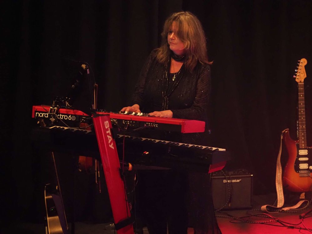 A musician plays a red and black keyboard setup on stage, lit warmly against a dark backdrop, with guitars and music equipment positioned around them.