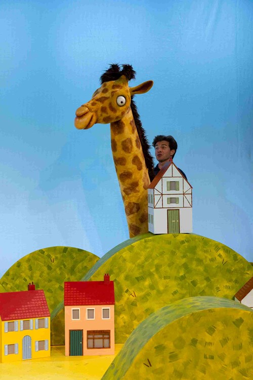 A giraffe puppet towers over the set of a small town. 