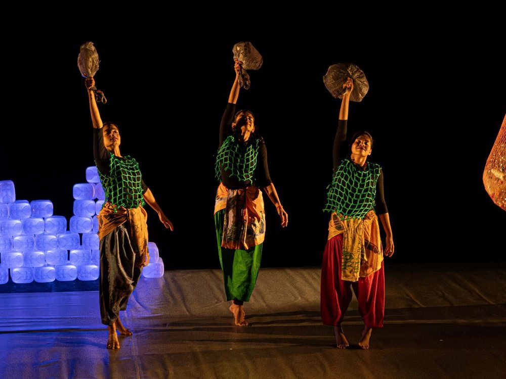 Three dancers perform on a dark stage, each holding a plastic object aloft. They wear colourful costumes with green netted overlays, while glowing stacks of plastic containers and a hanging net filled with plastic waste form part of the set.