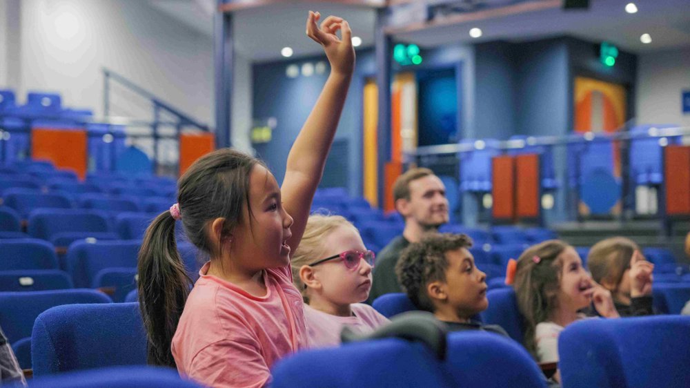 5 children and an adult sat in rows of seats looking up toward a stage. A child in the foreground has brown hair in a ponytail has raised her hand.