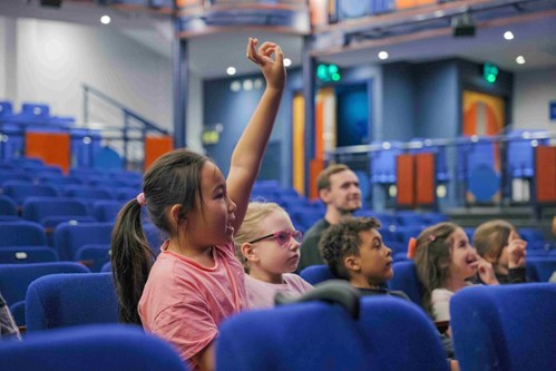 5 children and an adult sat in rows of seats looking up toward a stage. A child in the foreground has brown hair in a ponytail has raised her hand.