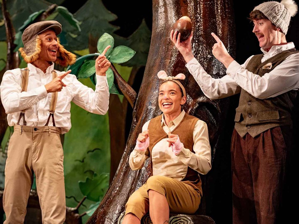 Three performers in woodland costumes gather around a tree. One actor holds up an acorn while another points at it, as a mouse-like character sits smiling beneath them.