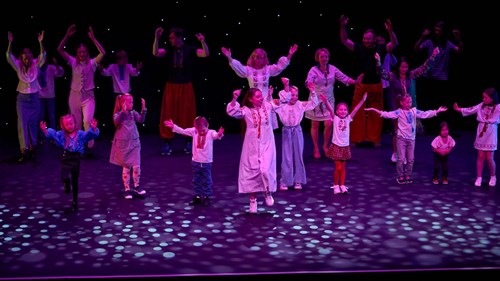 A diverse group of children and adults wearing traditional embroidered clothing perform a choreographed routine on a theatre stage. The background is black with star-like lights, and the performers raise their arms joyfully under purple and blue stage lightin