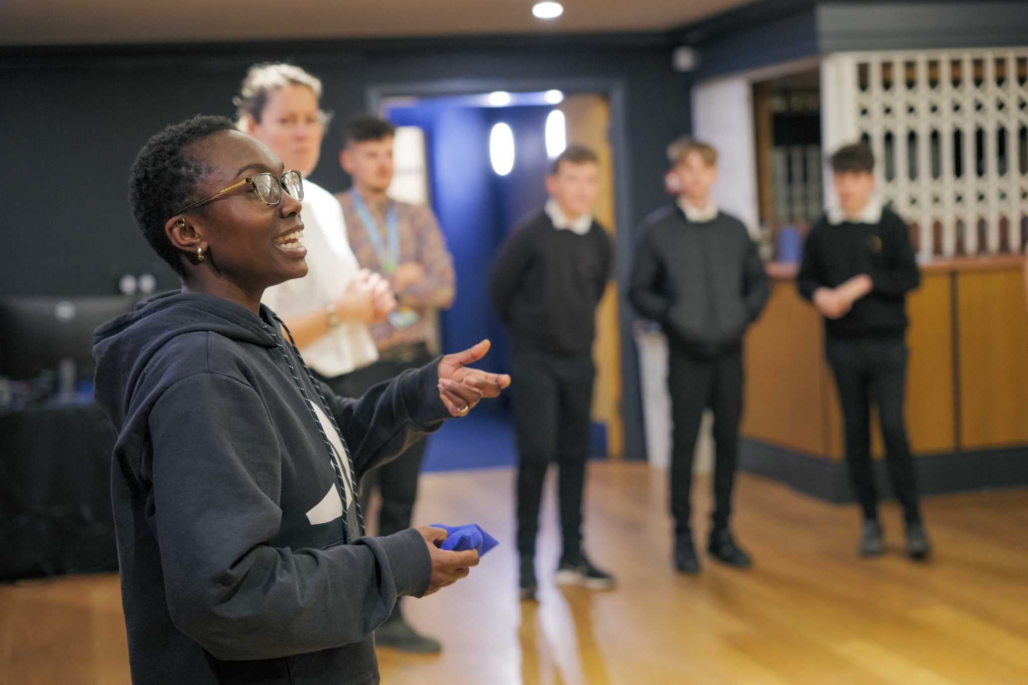 A theatre practitioner speaking to a small group of students during a workshop, with students standing and listening in the background.