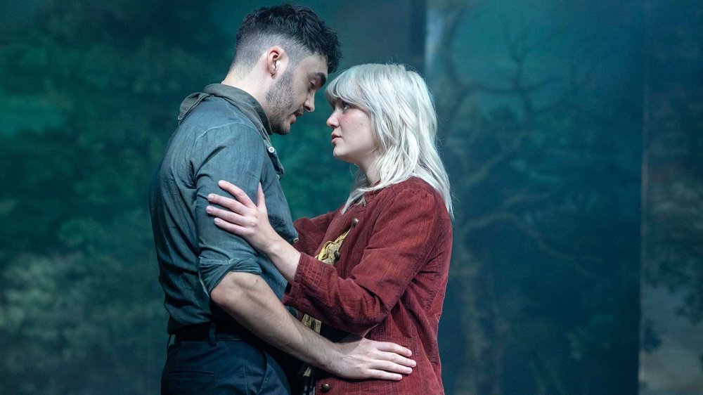 Two people embrace and gaze at each other on a forest-themed stage, creating an intimate, emotional moment.