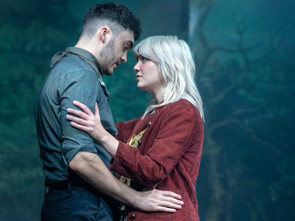 Two people embrace and gaze at each other on a forest-themed stage, creating an intimate, emotional moment.