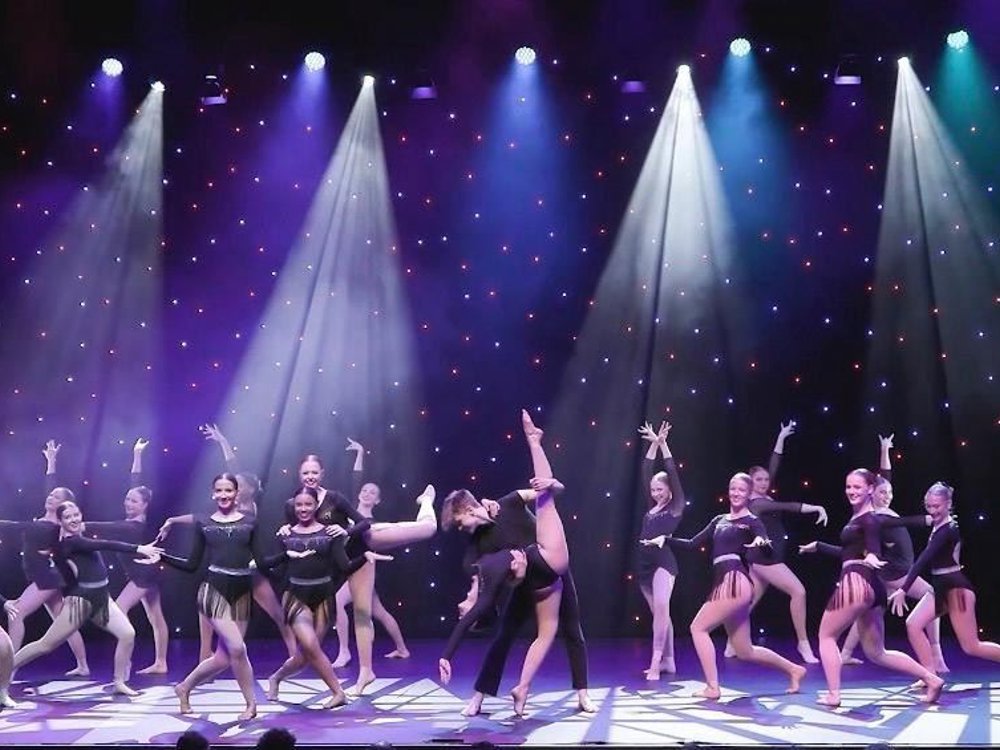 A large group of dancers perform on stage under bright spotlights, wearing coordinated dark costumes. They strike expressive poses and lifts against a starry backdrop, creating a dynamic, high-energy performance scene.