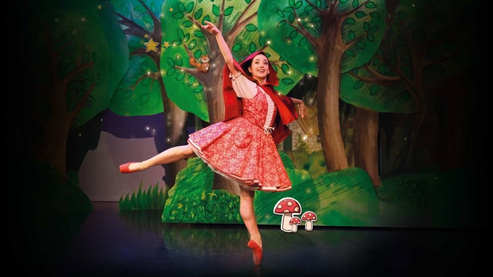 A ballet dancer dressed as Little Red Riding Hood performs a graceful pose on stage, surrounded by a colourful forest backdrop with illustrated trees, stars, and toadstools.